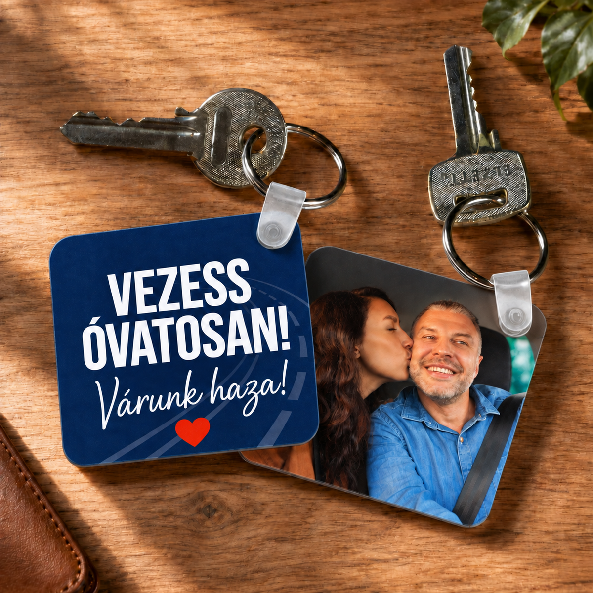 Kétoldalas műanyag fényképes kulcstartó párfotóval és sötétkék „Vezess óvatosan! Várunk haza!” grafikával, fa asztalon.