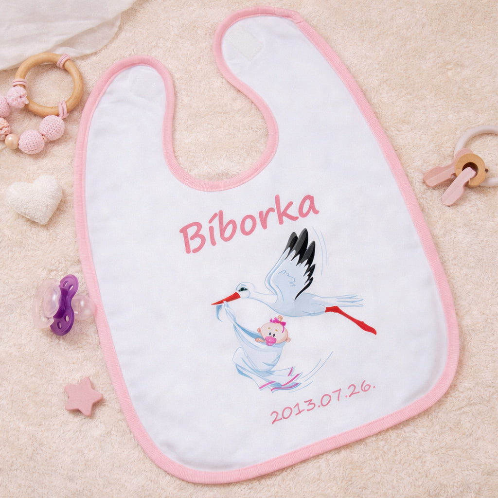Egyedi gólyás baba előke rózsaszín szegéllyel, cserélhető keresztnévvel és születési dátummal, gólya grafikával és újszülött babával, bézs háttéren esztétikusan elrendezve.