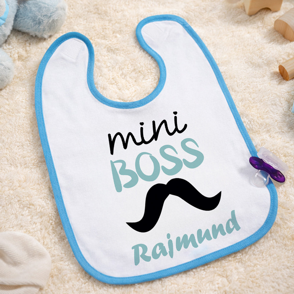Egyedi „mini BOSS” feliratú baba előke kék szegéllyel, fekete bajusz grafikával és cserélhető keresztnévvel, bézs háttéren esztétikusan elrendezve.
