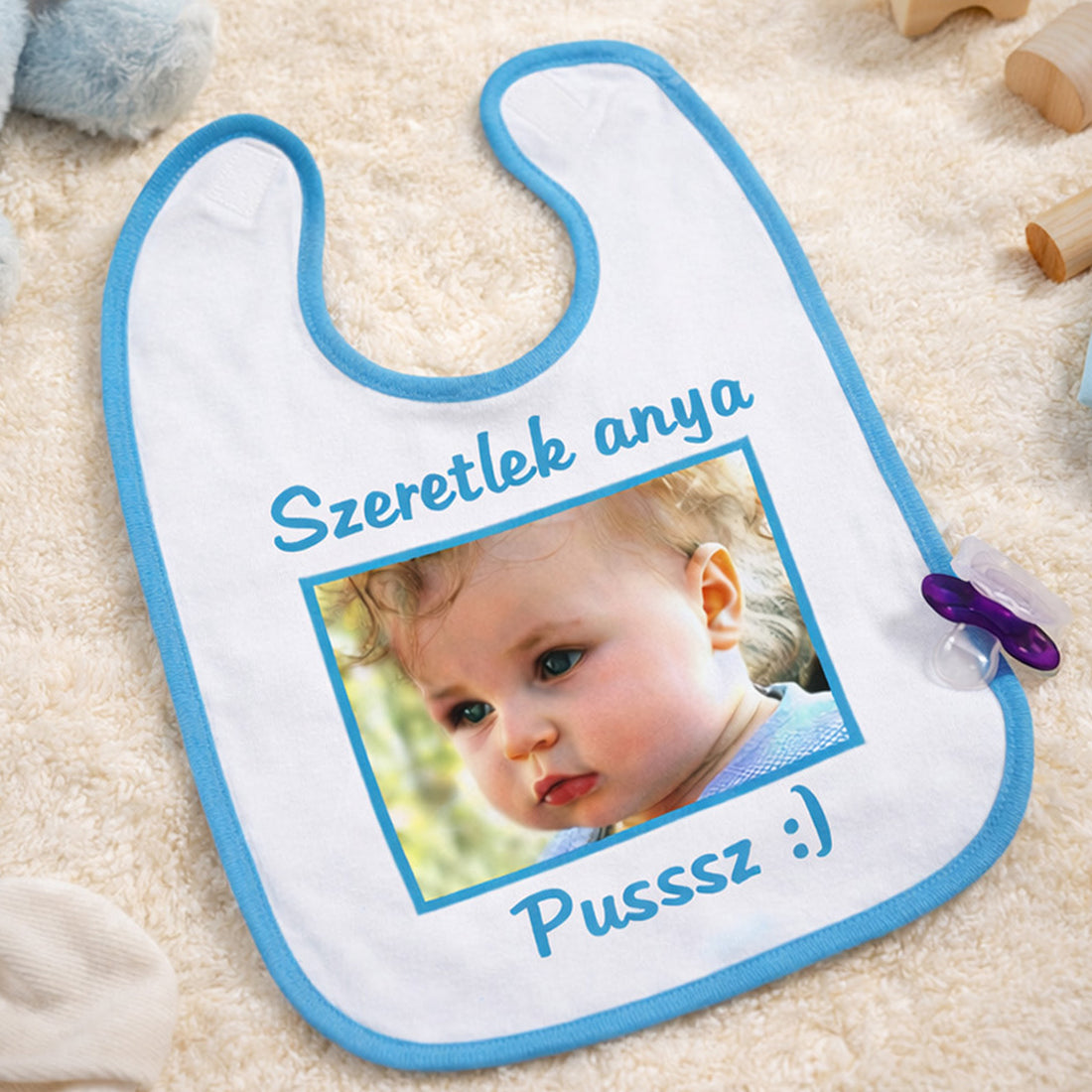 Egyedi fényképes baba előke kék szegéllyel, cserélhető felirattal és fotóval, bézs háttéren elhelyezve.
