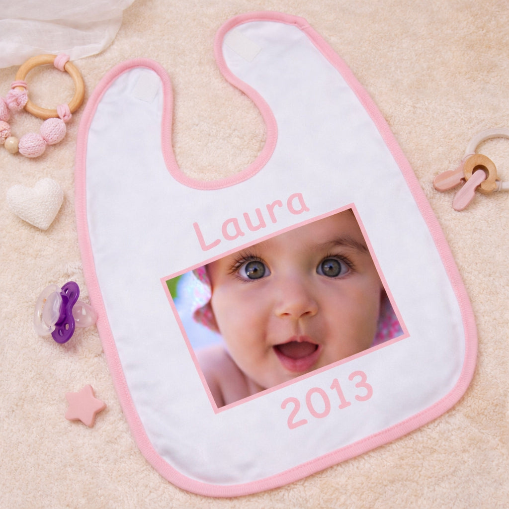 Egyedi fényképes baba előke rózsaszín szegéllyel, cserélhető névvel és évszámmal, középen beilleszthető fotóval, bézs háttéren esztétikusan elrendezve.