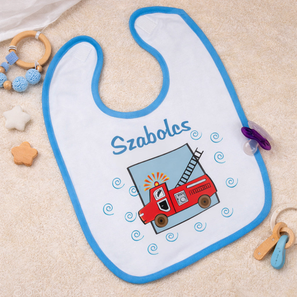 Egyedi tűzoltó mintás baba előke kék szegéllyel, piros tűzoltóautó grafikával és cserélhető keresztnévvel, bézs háttéren esztétikusan elrendezve.