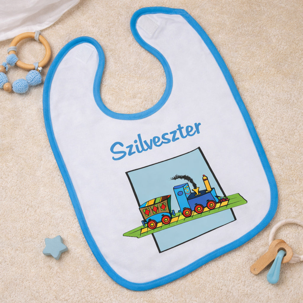 Egyedi vonatos baba előke kék szegéllyel, színes vonat grafikával és cserélhető keresztnévvel, bézs háttéren esztétikusan elrendezett babakellékekkel.